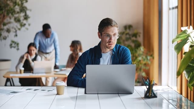 Man in glasses working on a laptop at a desk with a coffee cup, while three people collaborate at a table in the background.